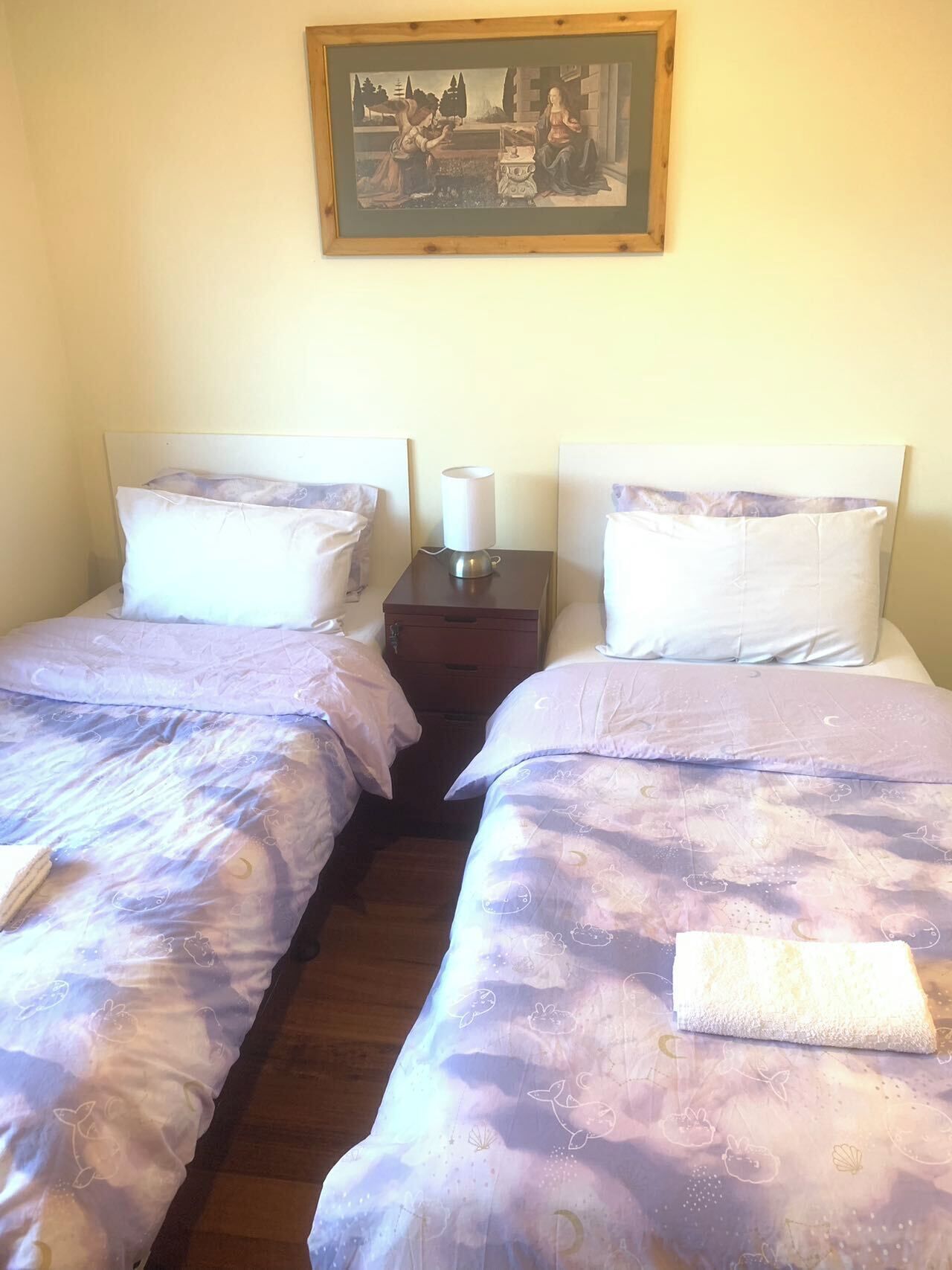 A hotel room with two single beds, a nightstand with a lamp, and a framed artwork above the beds.