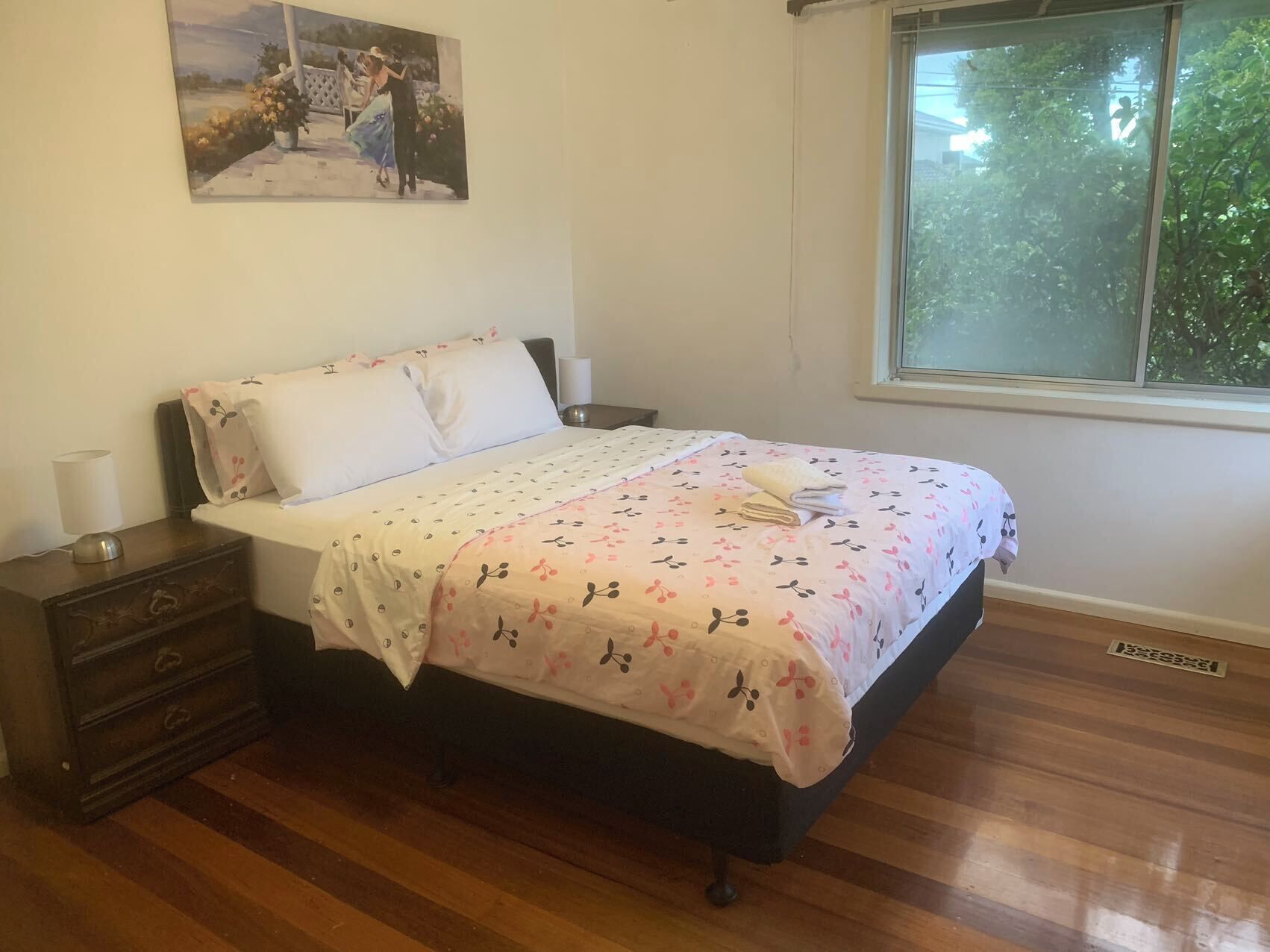 A bedroom with a wooden floor, a bed with a floral bedspread, a nightstand with a lamp, and a window with a view of greenery.