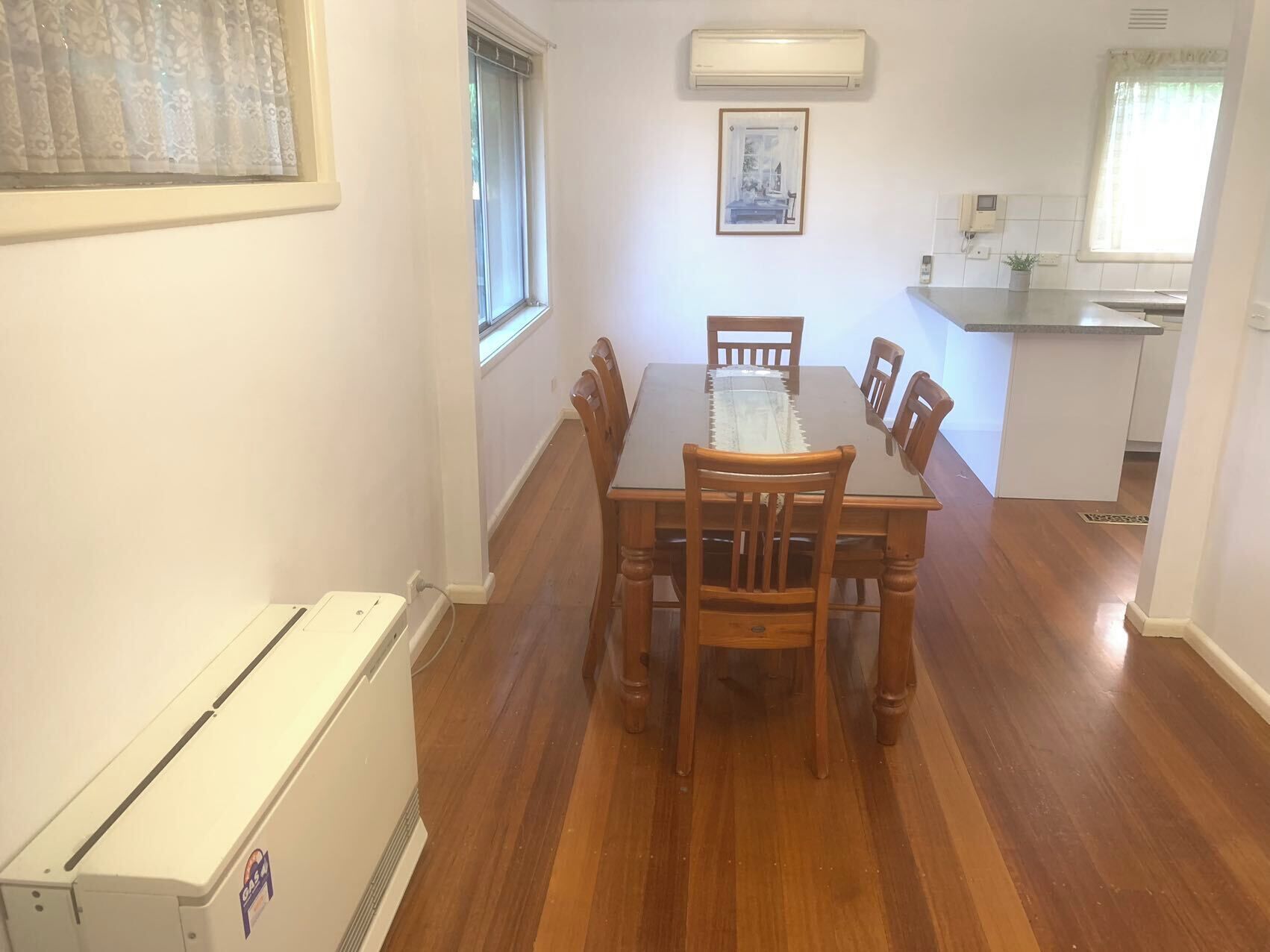A dining area with a table and chairs, a window, an air conditioner, and a kitchen in the background.