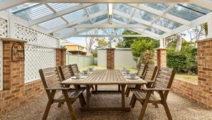 An outdoor patio with a wooden table and chairs, surrounded by brick walls and a glass roof structure.