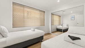 A modern bedroom with two beds, large windows with blinds, a mirror, and a small artwork on the wall.