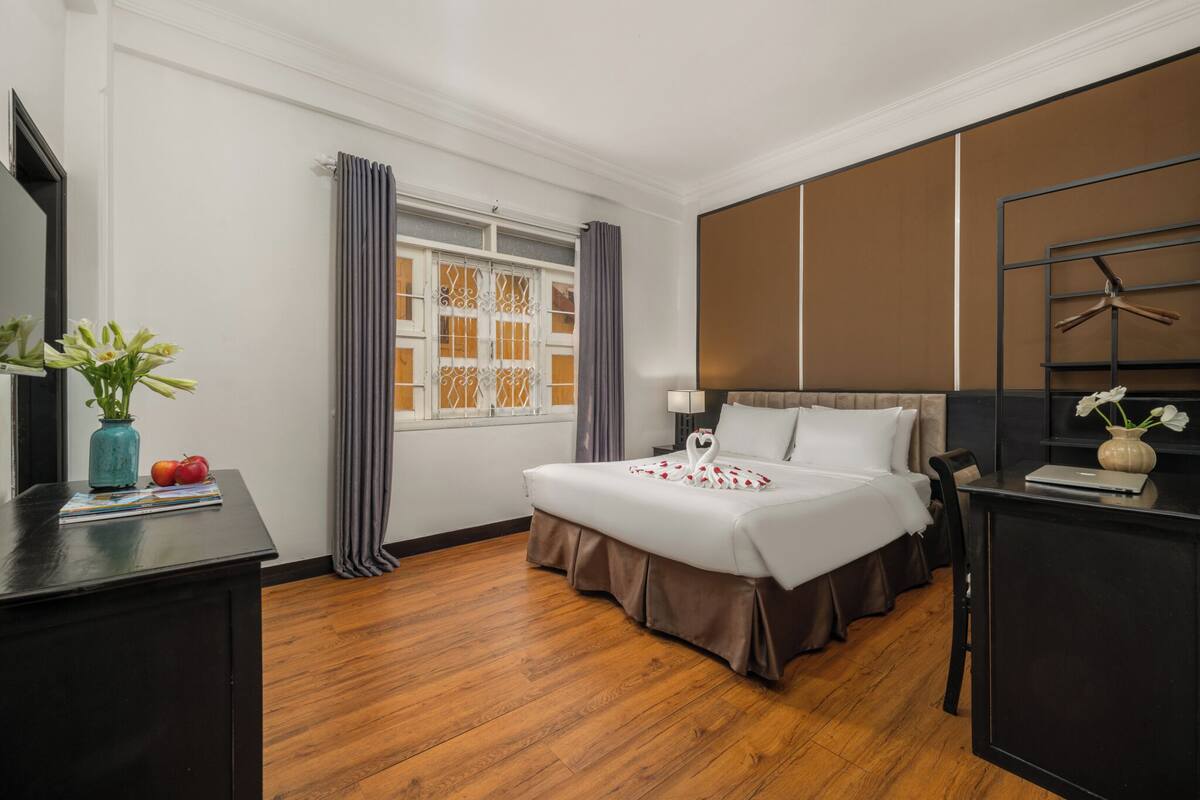 A hotel room with a bed, a desk, a window with curtains, a wall-mounted TV, and a vase with flowers.