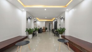 Uma área de recepção moderna, com um balcão extenso, plantas em vasos e um espaço de estar com mesas redondas.