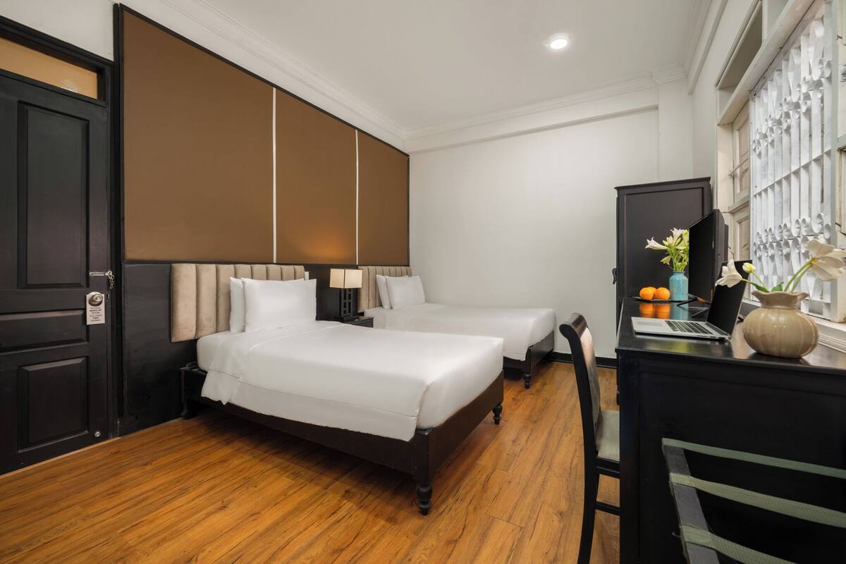 A hotel room with two beds, a desk with a laptop, a vase with flowers, and a window with a decorative grille.