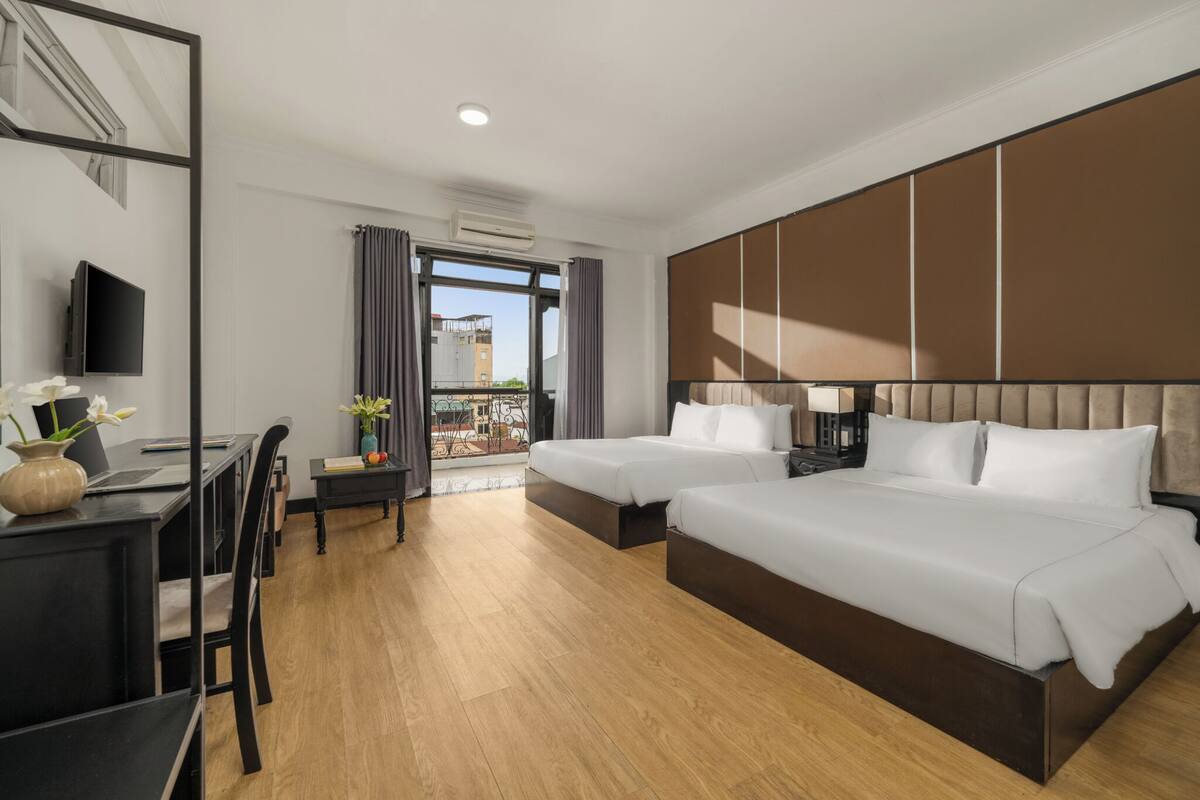 A hotel room with two beds, a desk with a chair, a TV, and a balcony with a view of buildings.