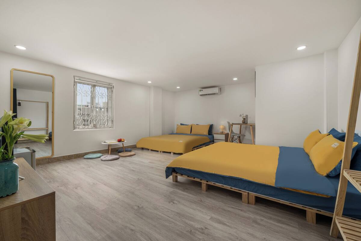 A modern hotel room with two beds, a wooden floor, a large mirror, a window with a view, and a small table with a plant.