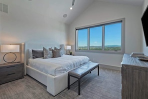 Une chambre à coucher avec un grand lit, des tables de chevet, un banc, une commode et une grande fenêtre donnant sur un beau paysage.