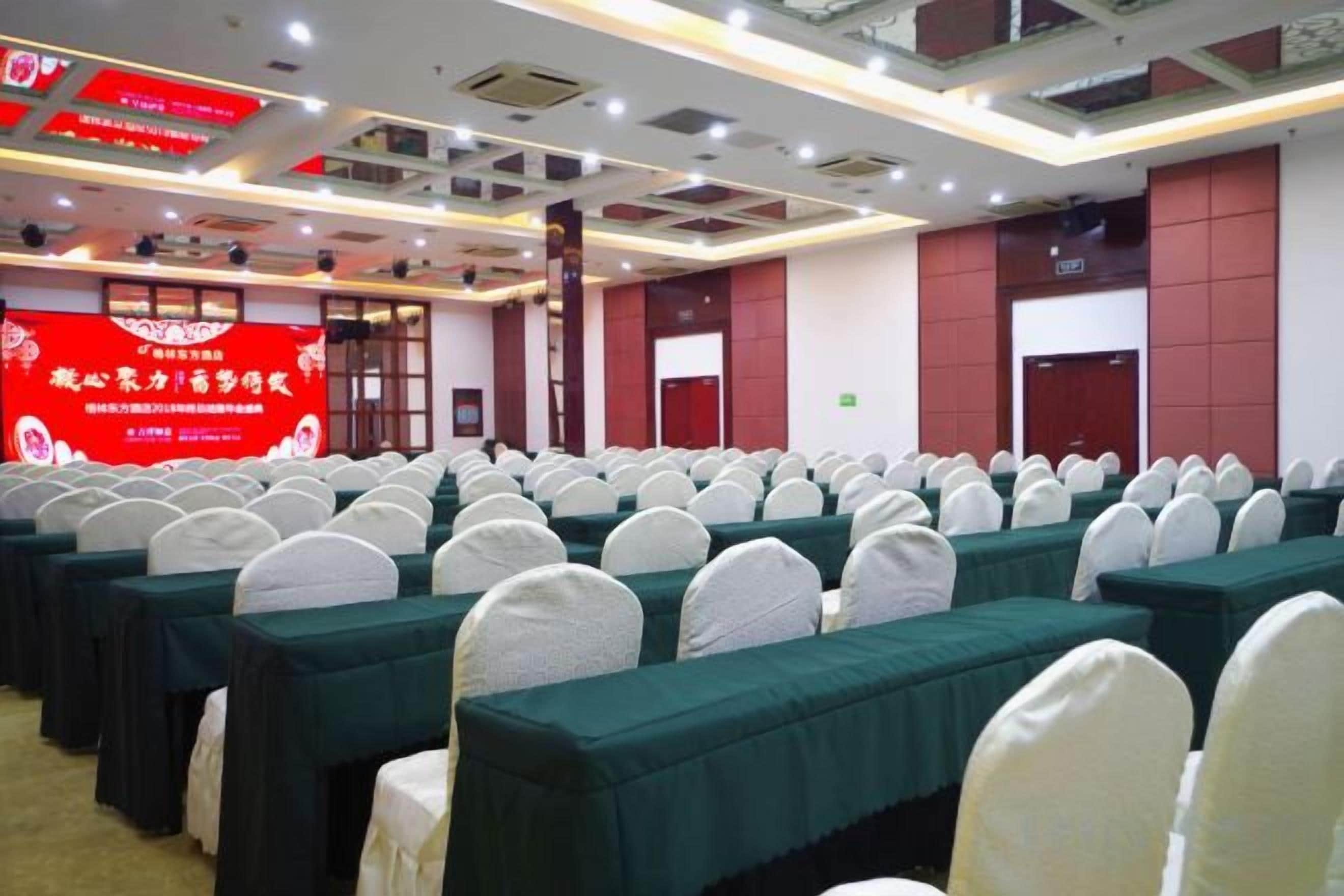 Un salón de conferencias con filas de mesas y sillas, un estandarte rojo y una puerta al fondo.