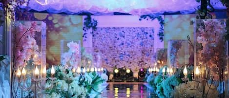 An indoor venue with a reflective floor, floral decorations, and a stage with a backdrop.