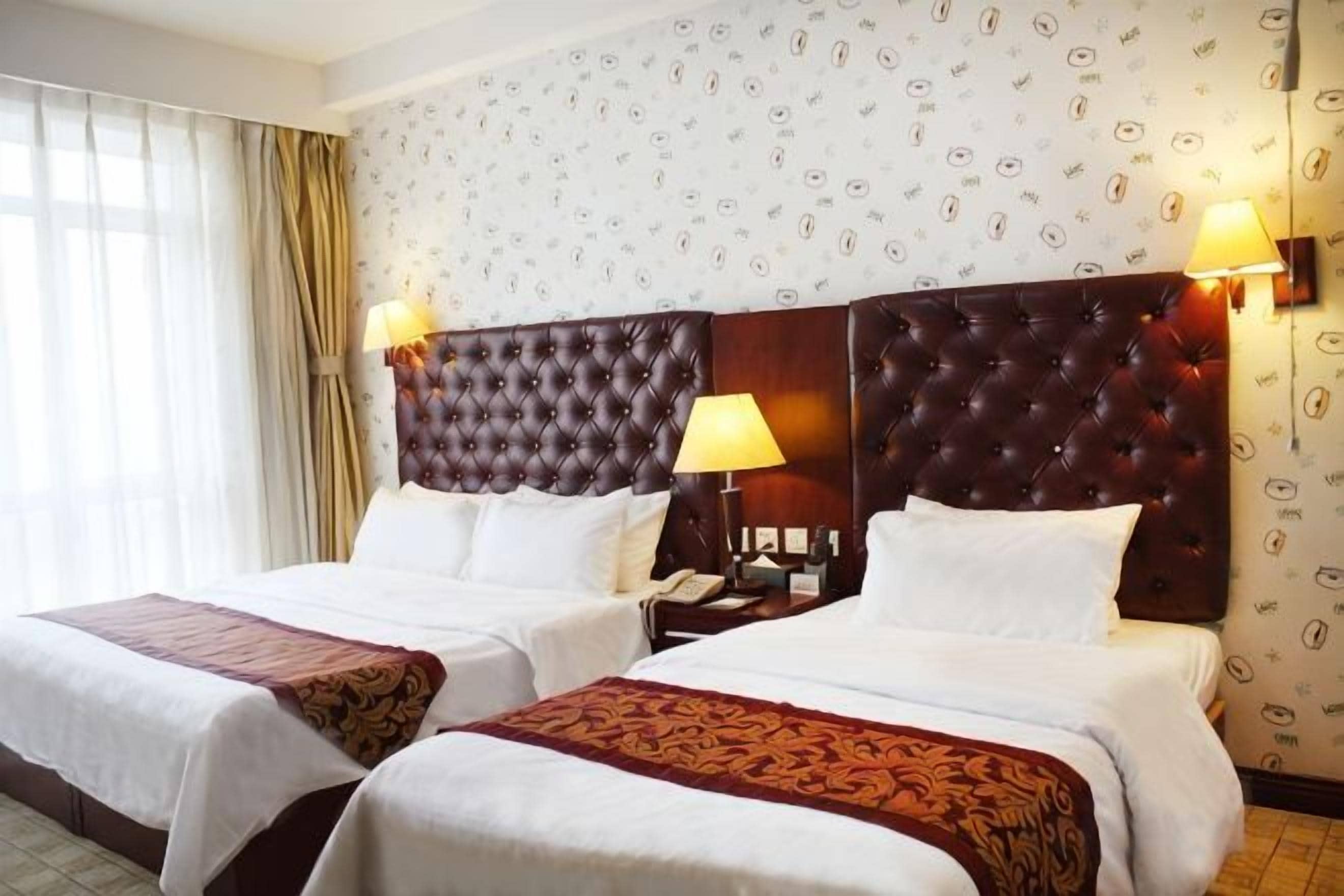 A hotel room with two beds, a large headboard, bedside lamps, and a patterned wallpaper.