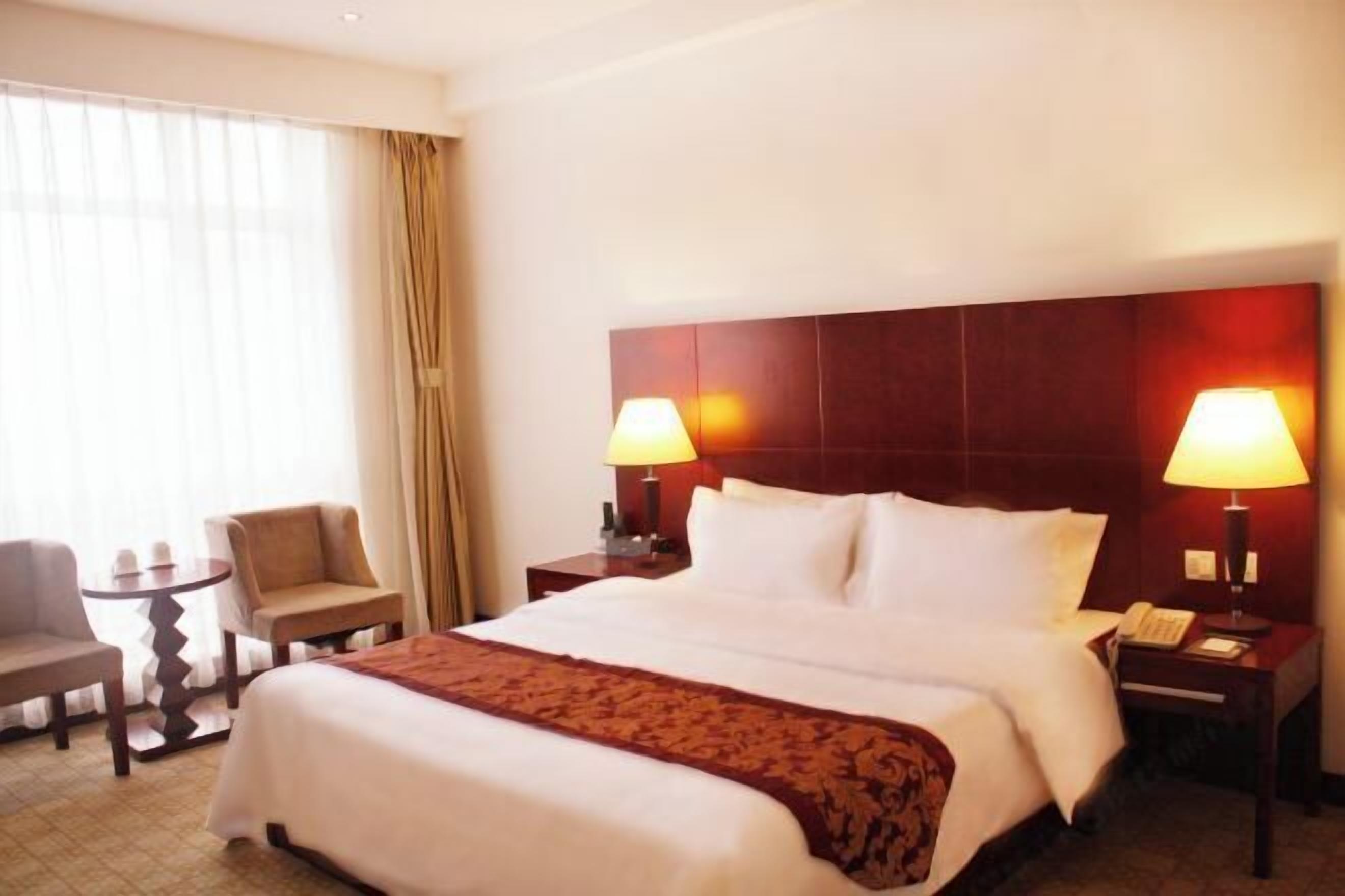 A hotel room with a large bed, two bedside lamps, a small table, a chair, and a window with curtains.