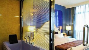 Habitación de hotel con una bañera grande, una cama con cabecera blanca, una mesita de noche con lámpara y vistas a una escalera.