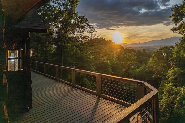 A wooden deck with a railing overlooking a forested landscape during sunset.