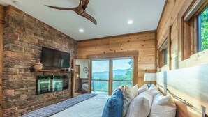 A bedroom with a stone wall, wooden ceiling, a bed with pillows, a television, and a view of the outdoors. - Luxury Cabin: Lake view/access, Theater, Game room (Bryson City)