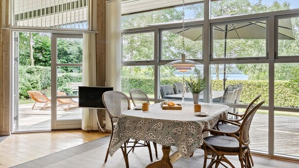 A dining area with a table set for four, surrounded by wicker chairs, large windows offering a view of greenery and a body of water, and a television on a stand. - Awesome home in Ry with WiFi (Ry)