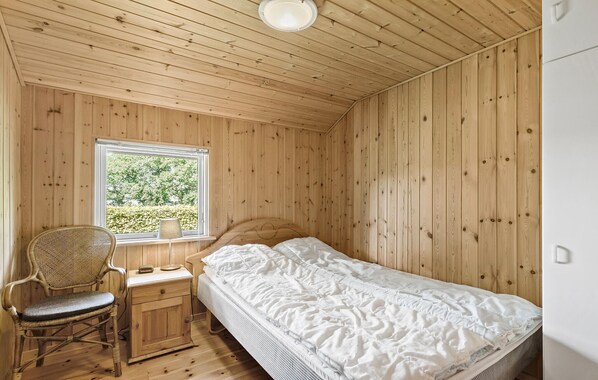 A wooden-paneled room with a bed, a chair, a nightstand, and a window.