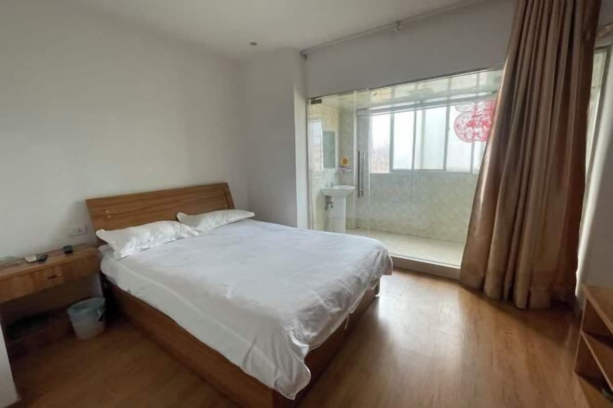 A bedroom with a wooden bed, a nightstand, a window with curtains, and a view of a balcony.