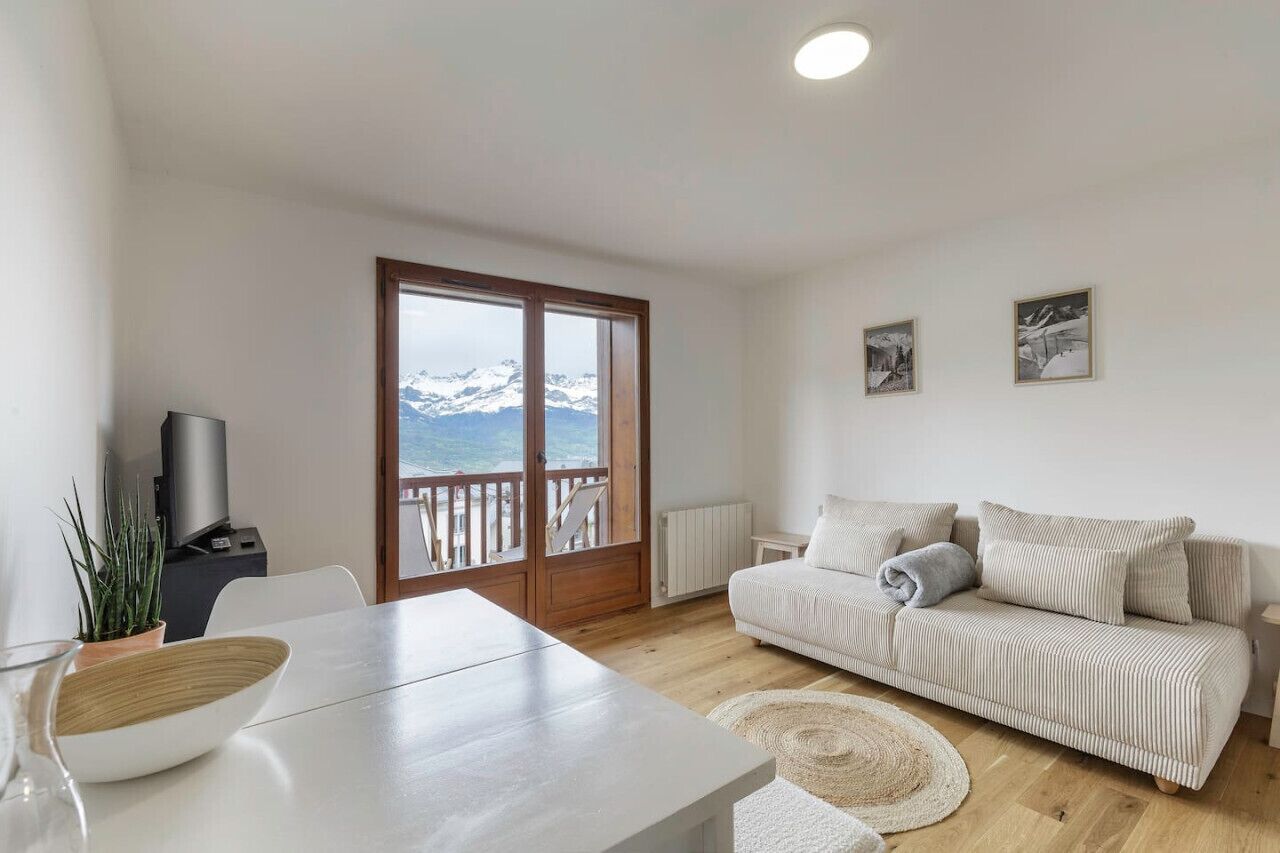 Un salon moderne avec un canapé, une table à manger et une vue sur les montagnes, visible à travers une porte vitrée.