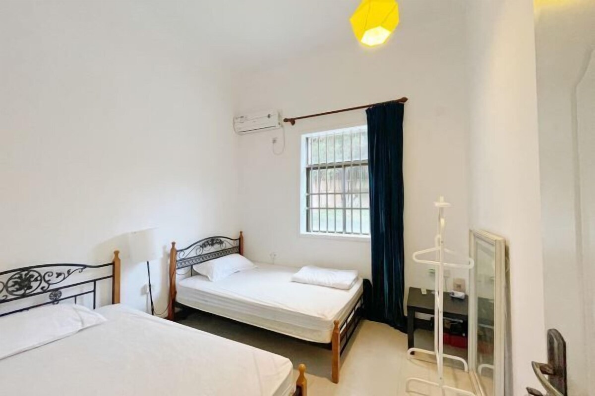 A small, clean room with two single beds, a window with curtains, a mirror, and a hanging light fixture.