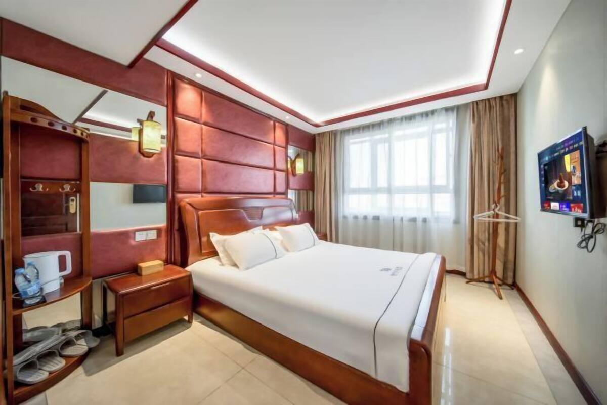 Une chambre d’hôtel avec un grand lit, une table de chevet avec une lampe, une armoire, une télévision et une fenêtre avec des rideaux.