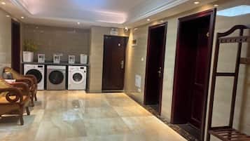 Un vestíbulo de hotel con piso de mármol, un área de descanso con sillas de madera, una fila de lavadoras y una puerta de color rojo oscuro.