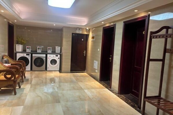 Un vestíbulo de hotel con piso de mármol, un área de descanso con sillas de madera, una fila de lavadoras y una puerta de color rojo oscuro.