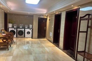 Un vestíbulo de hotel con piso de mármol, un área de descanso con sillas de madera, una fila de lavadoras y una puerta de color rojo oscuro.