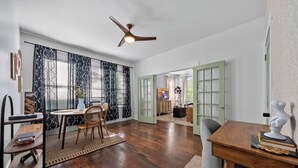 Un vaste espace de vie doté d’un plancher en bois, d’une table à manger avec des chaises, d’un ventilateur de plafond et d’une vue sur une autre pièce dont les portes sont vertes.
