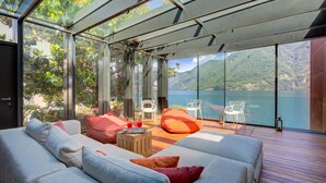A modern interior with a glass ceiling, a sofa set, a wooden table, and a view of a lake and mountains. - Maison Sur l'Eau (Paradiso)