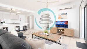 A modern living room with a sofa, coffee table, and TV. There is a kitchen area with white cabinets and a dining space with stools. - Zen Gem Pool Private Balcony Oceanview (Darwin)