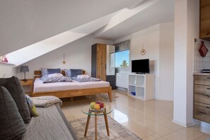 A modern bedroom with a large bed, a glass table with fruit, a TV, and a kitchen area.