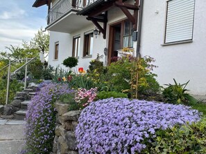 A white building with a balcony, stone steps, and a variety of flowers.
