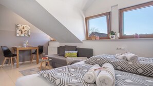 Ein Schlafzimmer mit Bett, zwei Kissen, einem Nachttisch, einem Sessel, einem kleinen Tisch, einem Fenster und einem Wandbild.