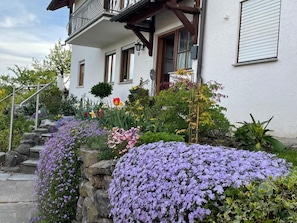 A white building with a balcony, stone steps, and a variety of flowers.
