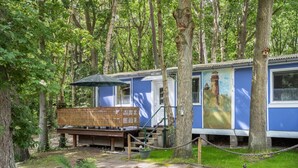 A blue mobile home with a deck, surrounded by trees, and a painting of a lighthouse on the side.