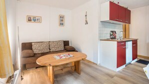 A compact living space with a sofa, a small wooden table, a kitchenette with red cabinets, and two framed pictures on the wall.