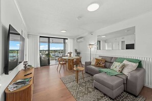 Un salon moderne avec un canapé gris, une table basse en bois, un coin repas avec une table et des chaises, et une grande fenêtre offrant une vue dégagée.