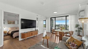 A modern living room with a flat-screen TV, a sofa, a dining area, and a balcony with a city view. - Stunning! 2Bed, 1Bath, 1Car, VIEWS ~ CBD (Brisbane City)