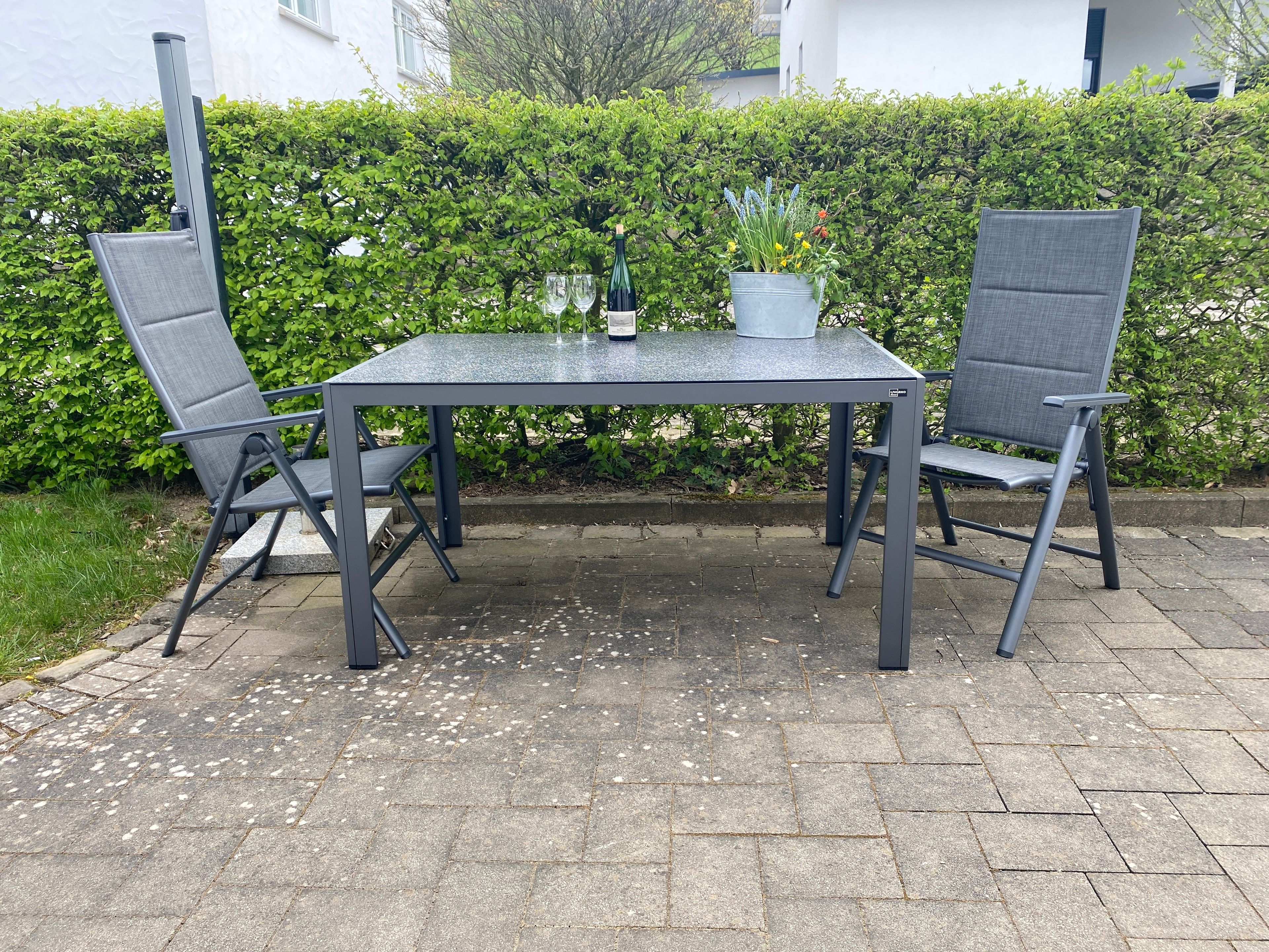 An outdoor patio with a table and two chairs, a potted plant, and a hedge in the background.