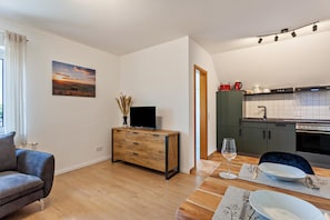 Un salon moderne doté d’un téléviseur à écran plat, d’une table à manger dressée avec des assiettes et des couverts, et d’une cuisine avec un dosseret carrelé et des appareils électroménagers.