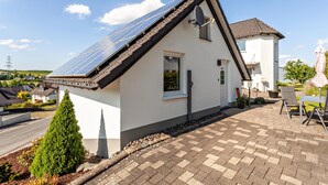 Une maison avec un toit en pente, des panneaux solaires, un petit jardin et une terrasse dallée.