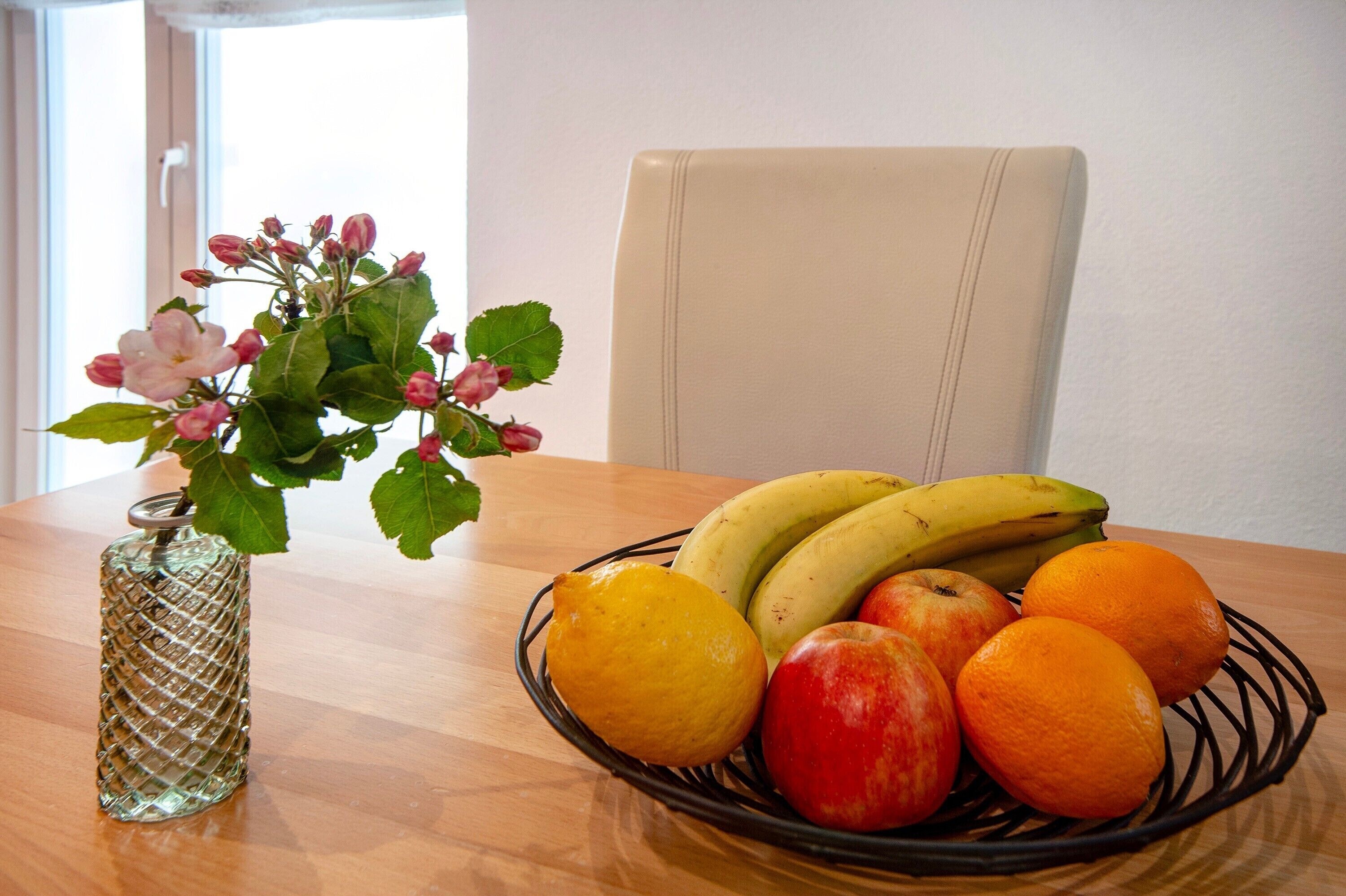 A dining table with a fruit bowl containing bananas, apples, and oranges, and a vase with flowers.