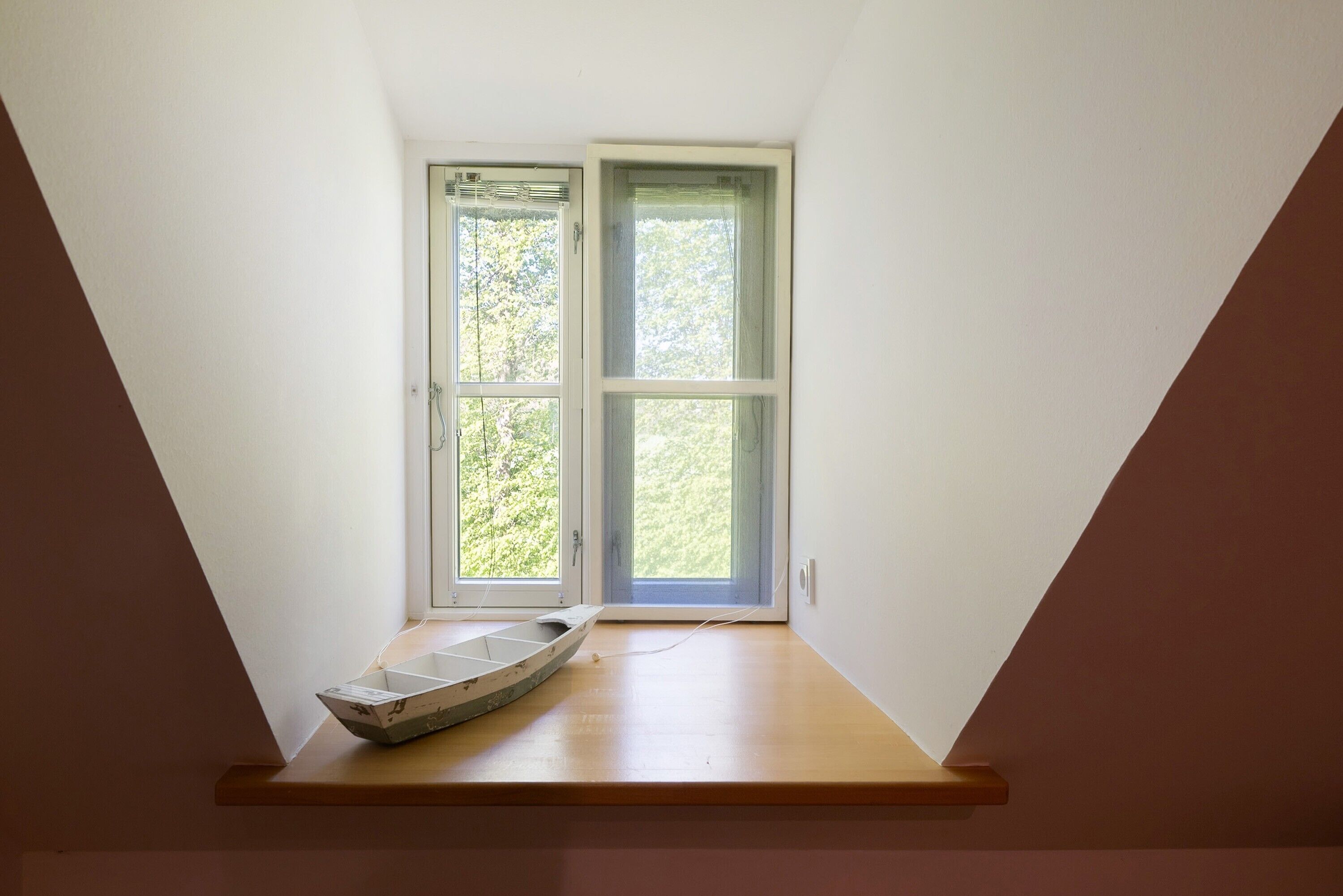 A room with a sloped ceiling, a small boat on a wooden ledge, and a window with a view of trees.