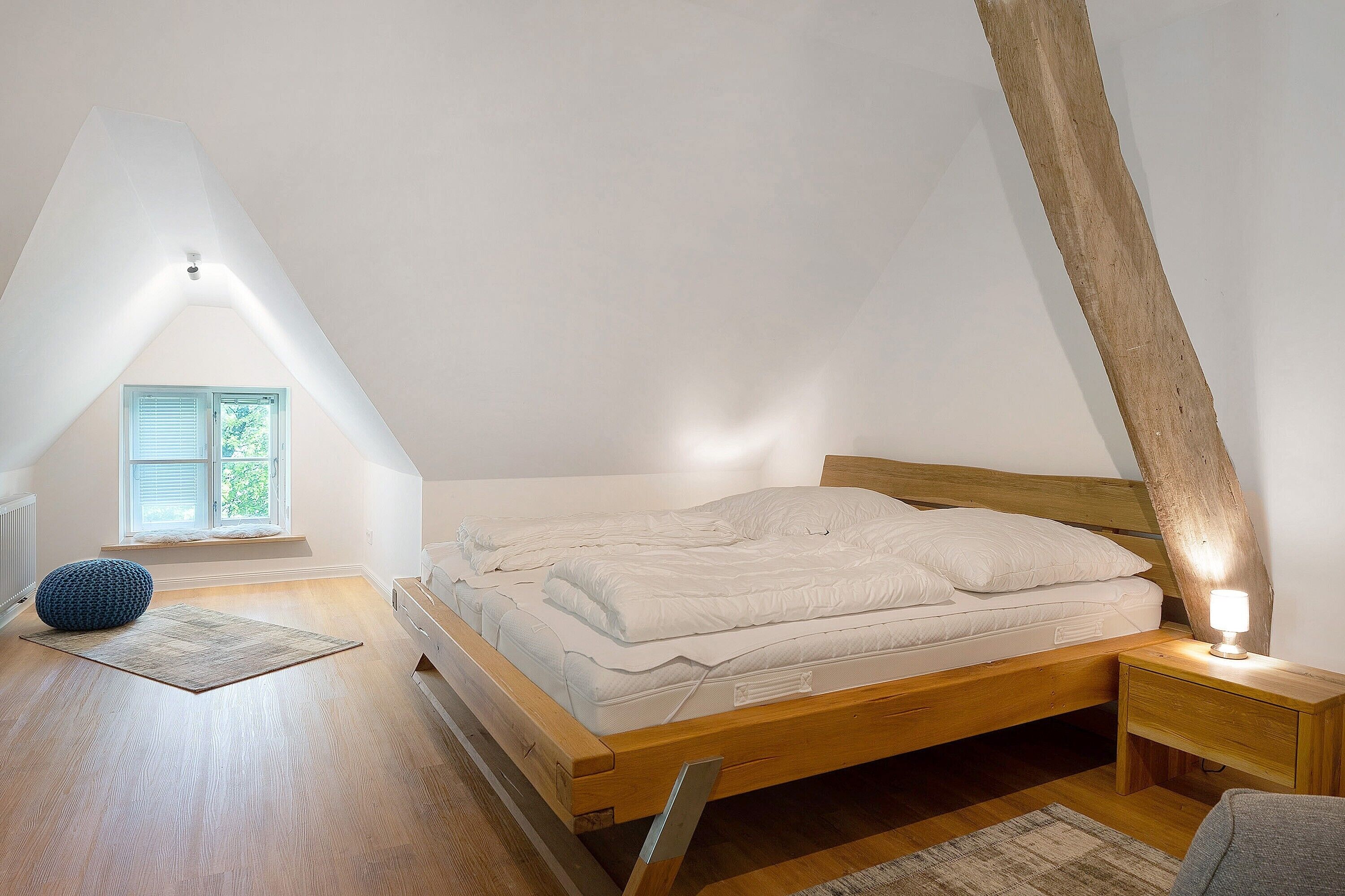 A bedroom with a sloped ceiling, a bed with white bedding, a bedside table with a lamp, and a window with shutters.