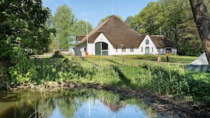 Une maison traditionnelle avec un toit de chaume, un étang au premier plan et une table de pique-nique à proximité.
