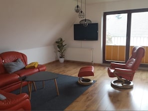 A living room with a red sofa, a red chair, a small table, a potted plant, and a TV mounted on the wall.