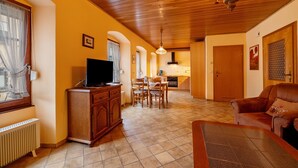 A living room with a television, wooden furniture, a dining area, and a kitchen in the background.