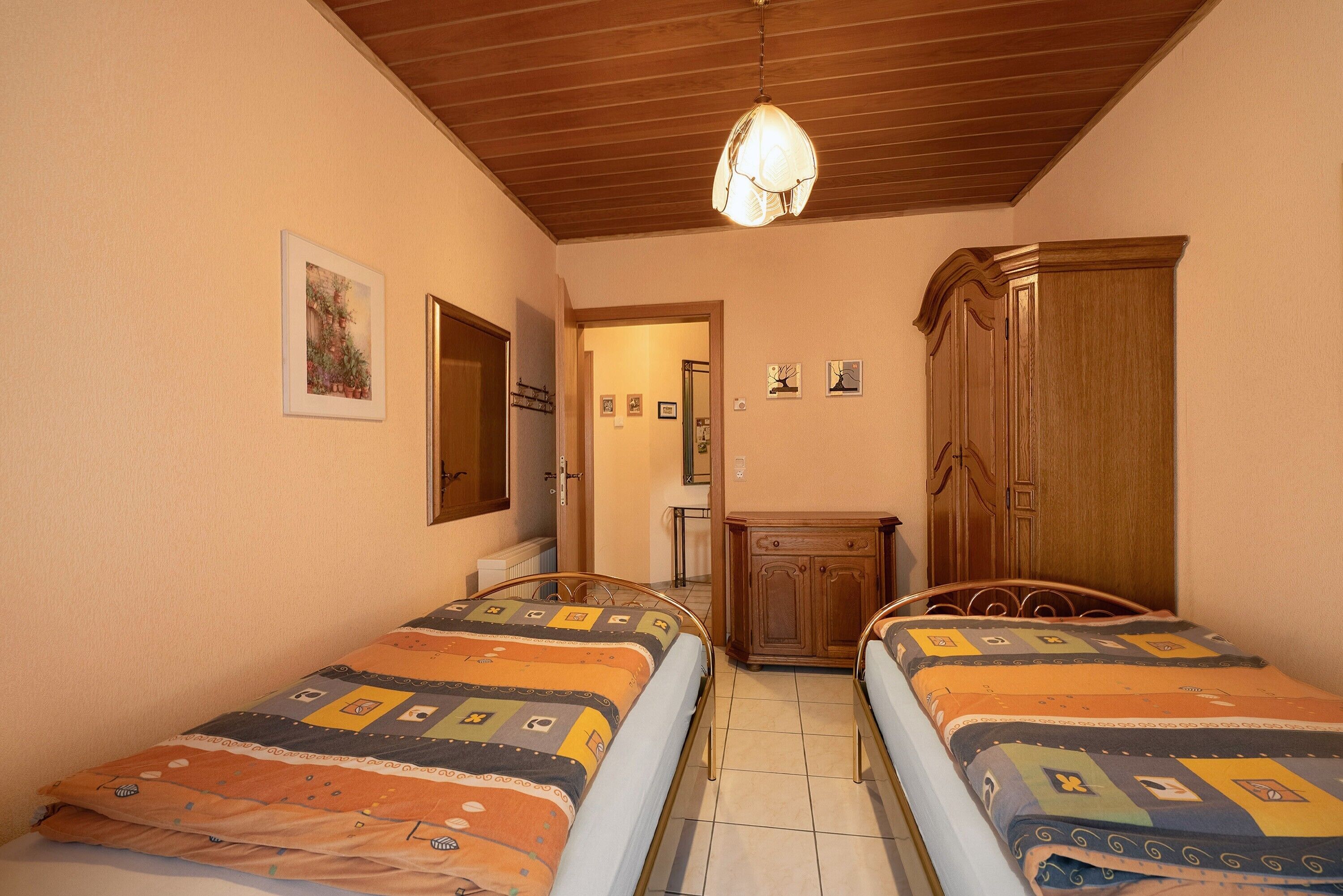Un dormitorio con dos camas, un armario de madera, una lámpara en el techo y una puerta que da a otra habitación.