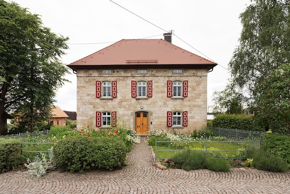 A two-story stone building with a red-tiled roof, a central entrance, and a paved driveway.