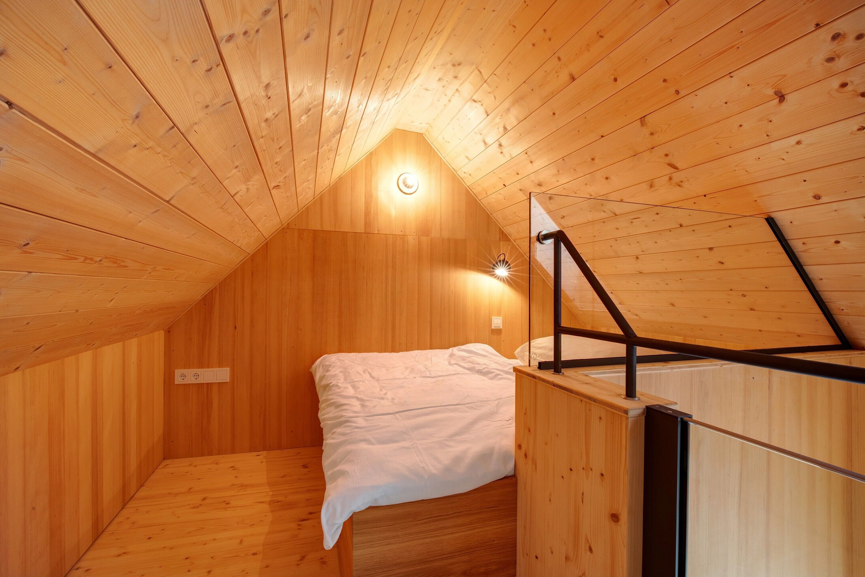 Un dormitorio bajo los tejados, pequeño y con paneles de madera, con una cama individual, techo inclinado y una lámpara.